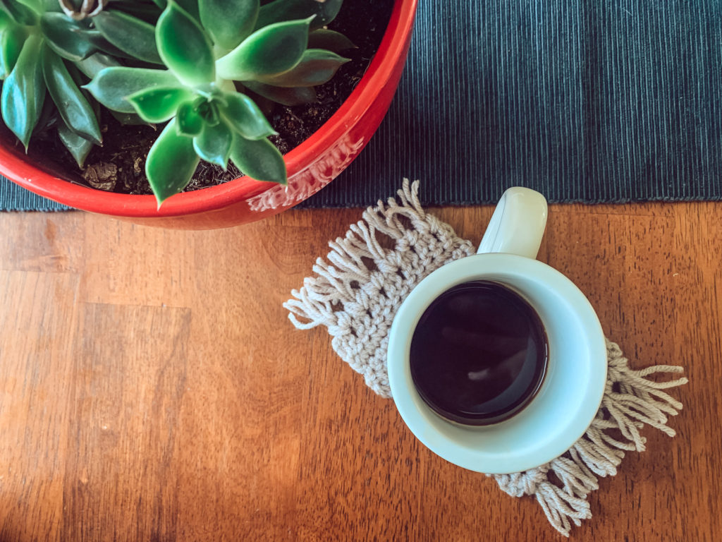 A mug sits on beige mug rug, a beginner crochet pattern, on a wood table along with a green plant and black table runner.