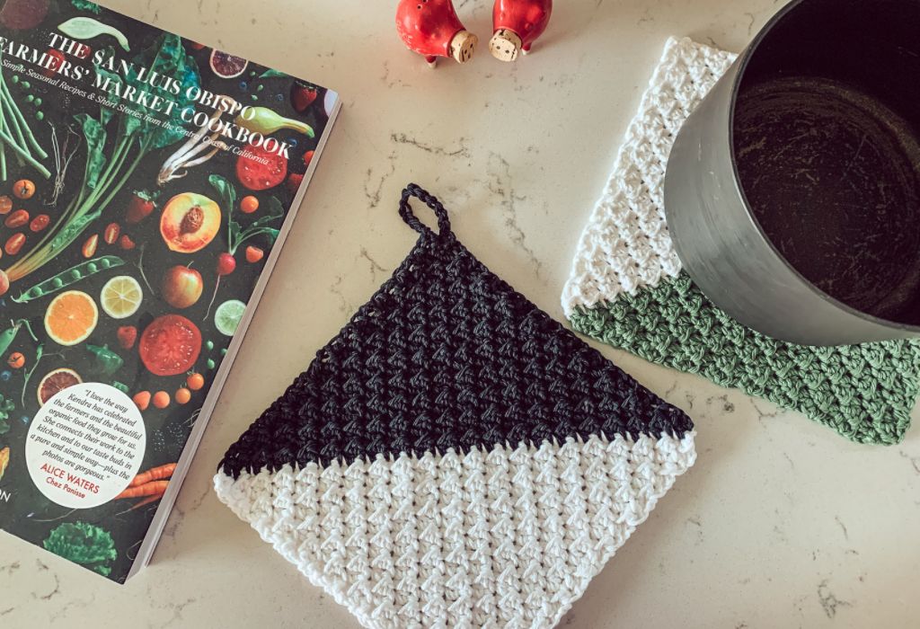 Black and white crochet pot holder made with the even moss stitch is on a marble counter, along with a cook book, pot, and red salt and pepper shakers