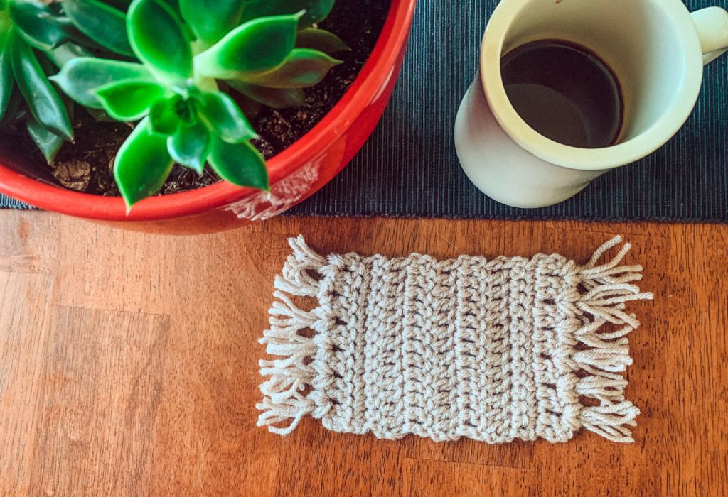 A beige mug rug, which is a beginner crochet pattern, is on a table next to a black table runner with a mug of coffee and a green plant