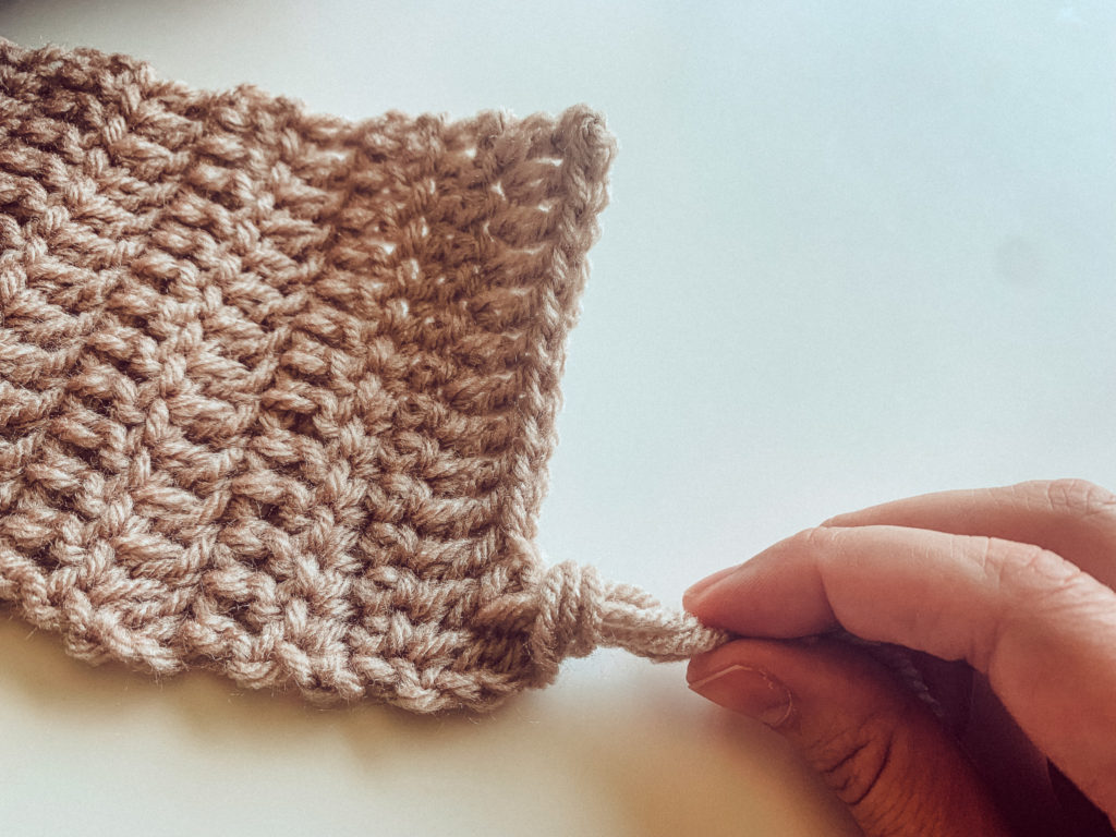 A hand is pulling  yarn through the end of a beige crochet mug rug to make fringe