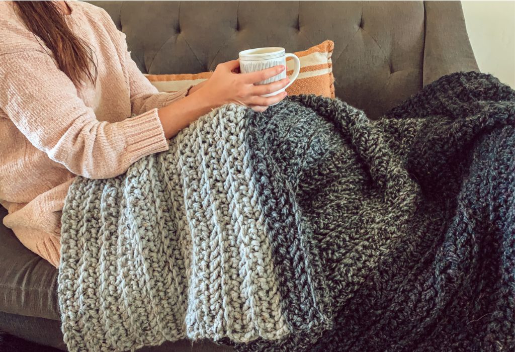 Woman sits on couch holding coffee with free modern crochet blanket pattern. The blanket is color-blocked and made with herringbone double crochet stitch.