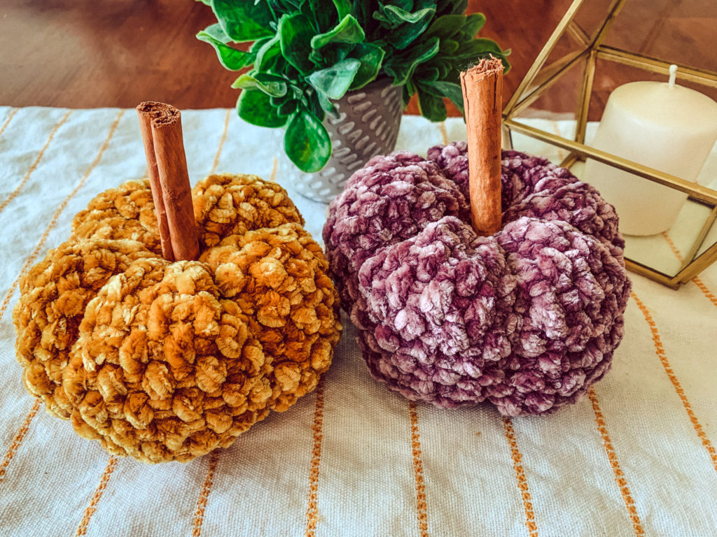 This image shows two velvet crochet pumpkins on a table runner, one mustard and one purple, with a candle and a potted plant in the background