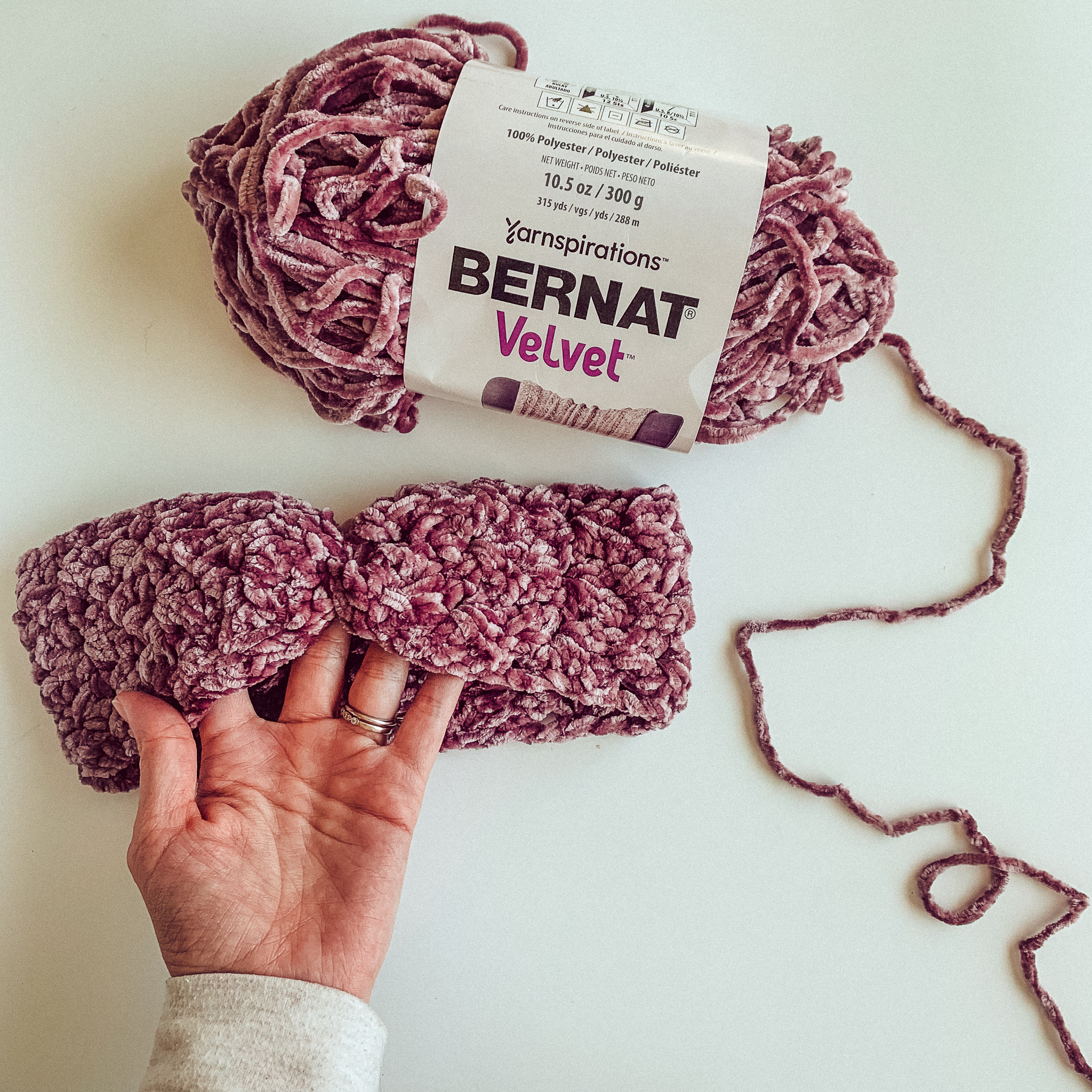 The photo shows a skein of purple velvet yarn, and a hand holding a crochet ear warmer made from same yarn. The background is white.