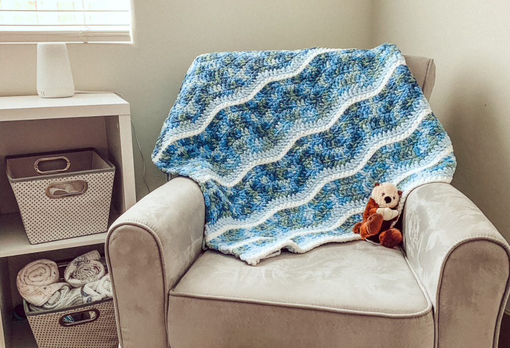 The image shows a crochet baby blanket made to look like ocean waves thrown over the back of a gray rocking chair. There is also a toy otter on the chair. The chair appears to be in a nursery with open white shelving next to it. The shelving has grey and white baskets in it. The blanket was made from a free crochet baby blanket pattern.