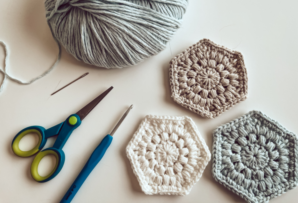 This photo shows the following items on a white background: a light blue ball of yarn, a darning needle, a pair of blue yarn scissors, a blue crochet hook, and three Hexagon shaped face scrubbies made from the Reusable Face Scrubbie free crochet pattern.  The face scrubbies are in the colors white, light blue, and tan.