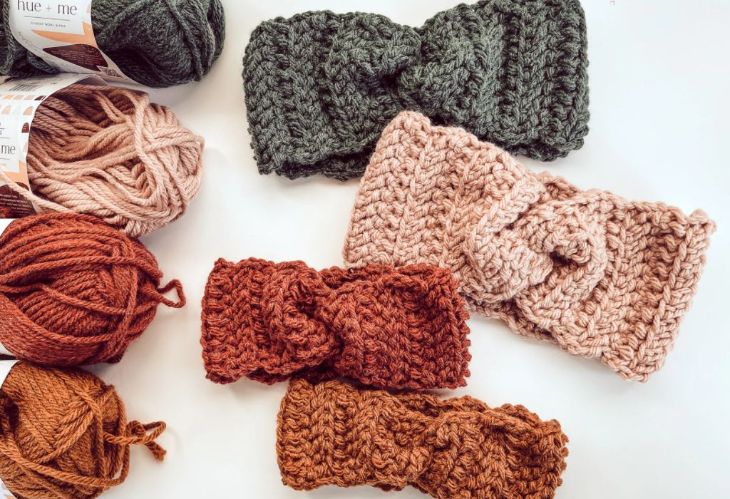 Four crochet twisted ear warmers are on a white surface with four skeins of yarn in matching colors. The yarn colors are dark green, beige, rust, and a dark gold.