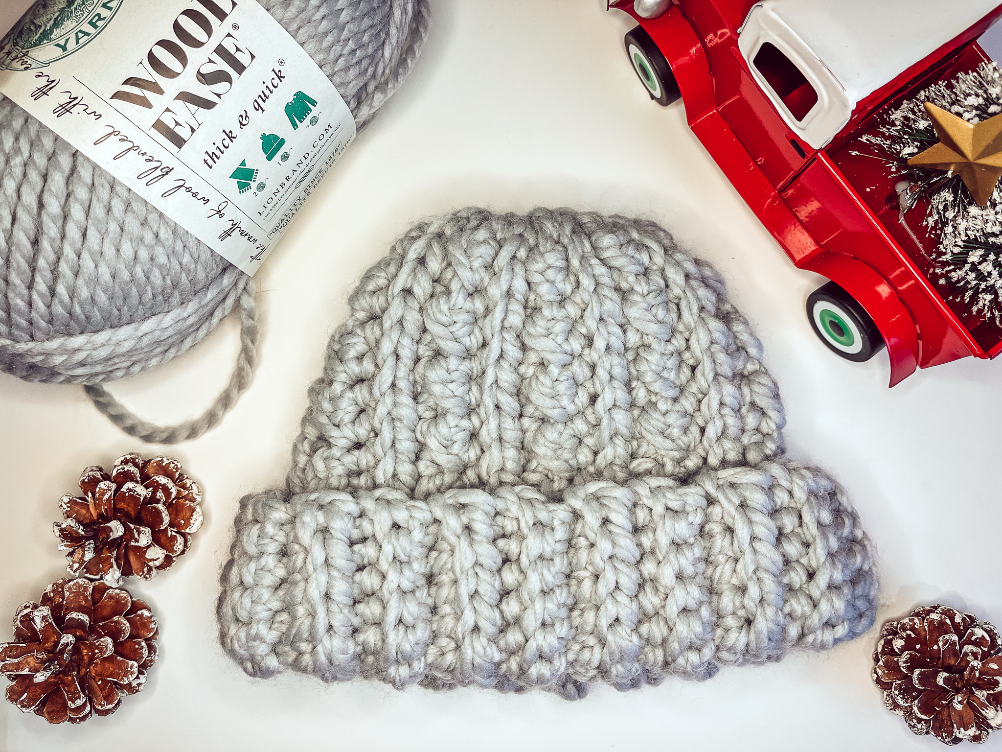 A gray, textured chunky crochet beanie is laying flat on a white background surrounded by pinecones, a toy red truck, and a ball of gray wool-ease quick and thick yarn.