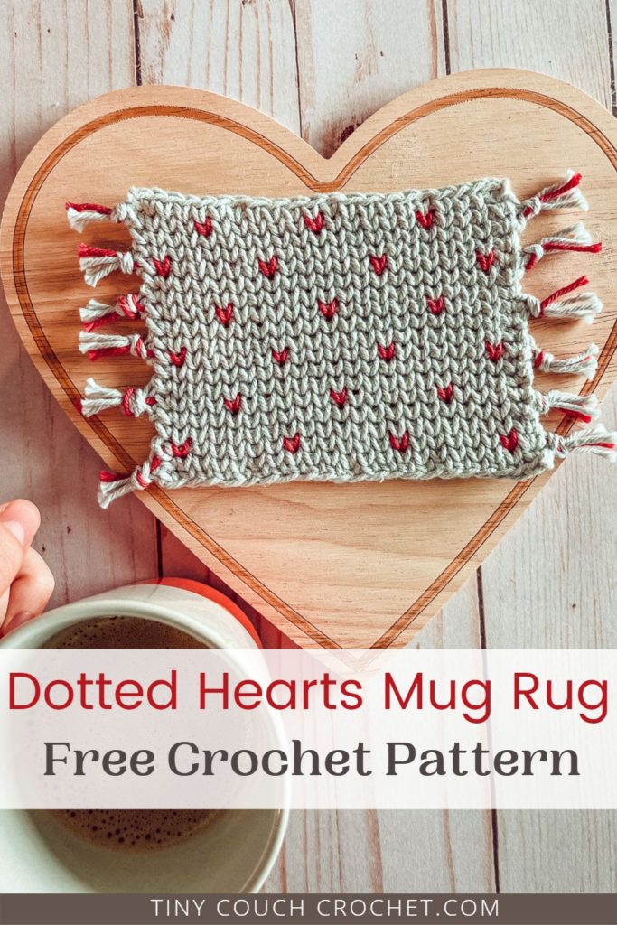 A gray coaster with pink dots in the shape of hearts is sitting on a large heart shaped wood board. A hand holding a mug of coffee is in the bottom corner. In the lower third of the image, text says "dotted hearts mug rug, free crochet pattern" and the very bottom of the image is text that says "tinycouchcrochet.com"