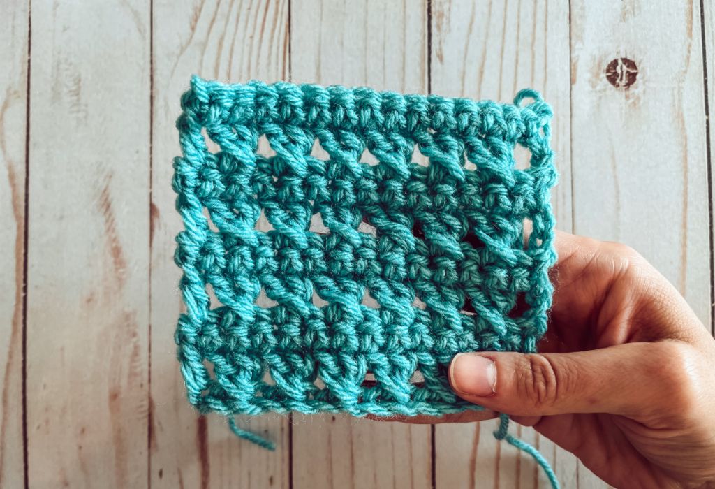 A blue crochet swatch of the crossed double crochet stitch is being held up by a hand. The background is whitewashed wood.