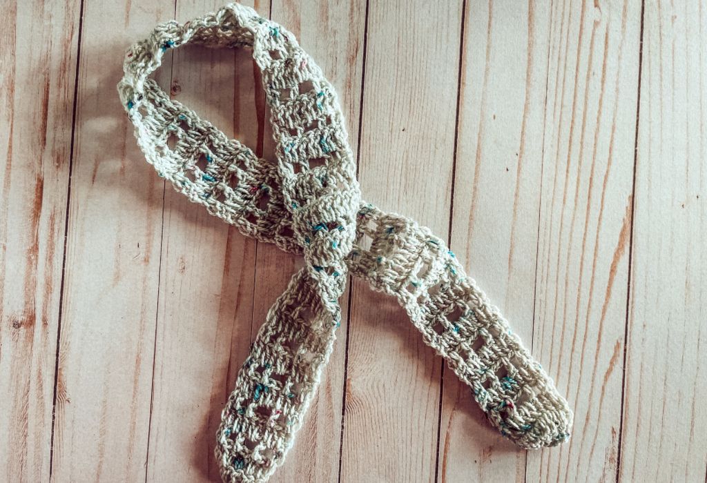 A crocheted, lacy hair scarf made with speckled yarn is laying on a wood background.