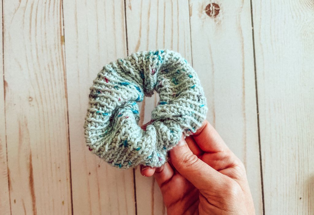 A hand is holding a blue-toned crochet scrunchie over a wood board background.