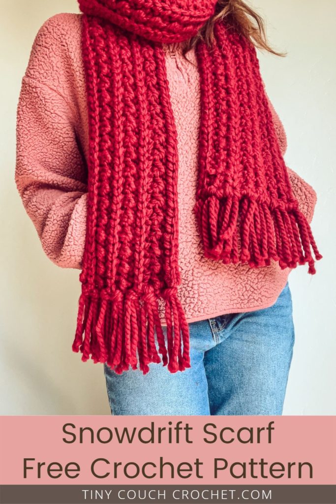 Photo of a woman wearing a pink fleece jacket and a chunky crochet scarf made with bulky red yarn. The bottom of the pin says "snowdrift scarf free crochet pattern tinycouchcrochet.com"