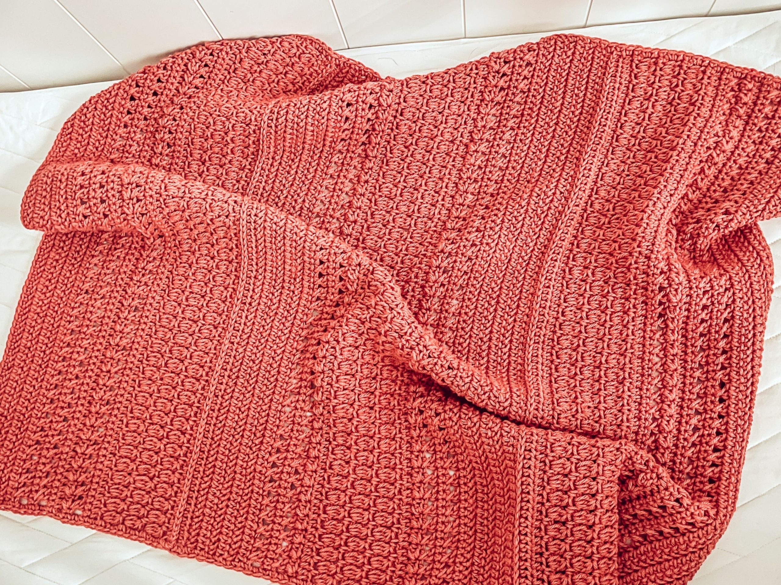 A close up of a pink stitch sampler crochet baby blanket is laying in a white crib.