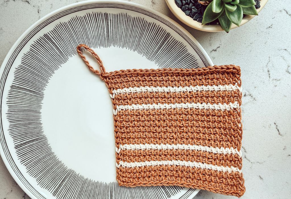 A Tunisian Crochet dishcloth that is mustard with white stripes is laying on a ceramic plate on top of a marble counter with a plant shown in the upper right corner. The discloth was made from a free beginner tunisian dishcloth pattern.
