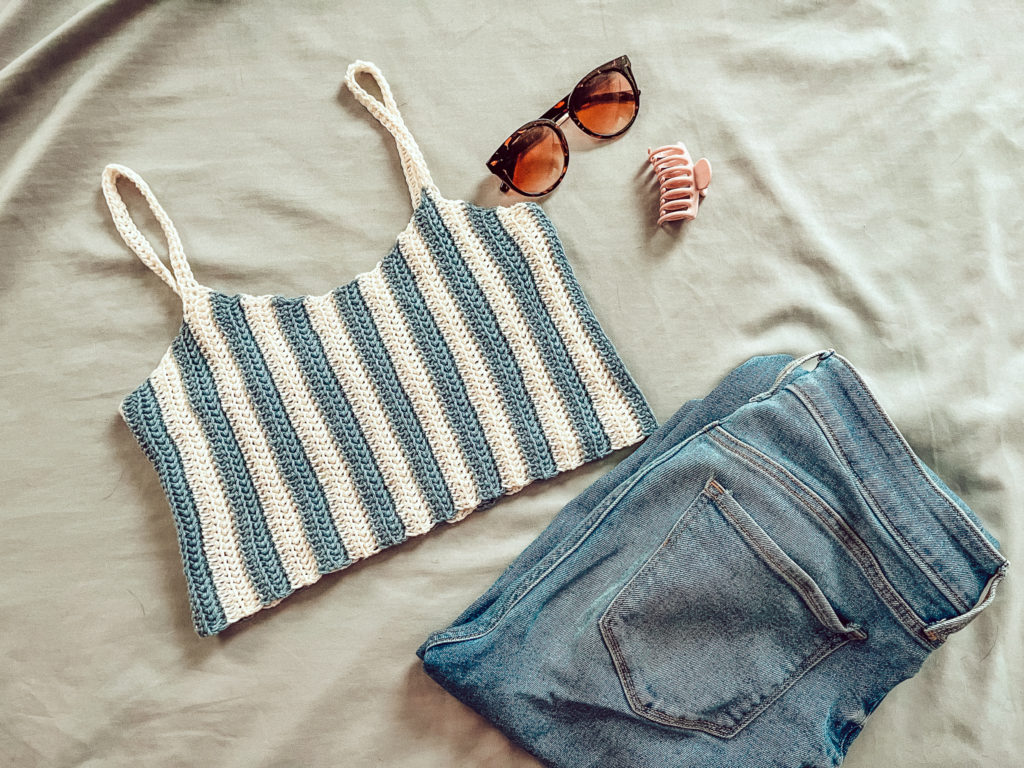 a blue and white striped crocheted crop top is on a bed with a pair of jeans, sunglasses and a hair clip.