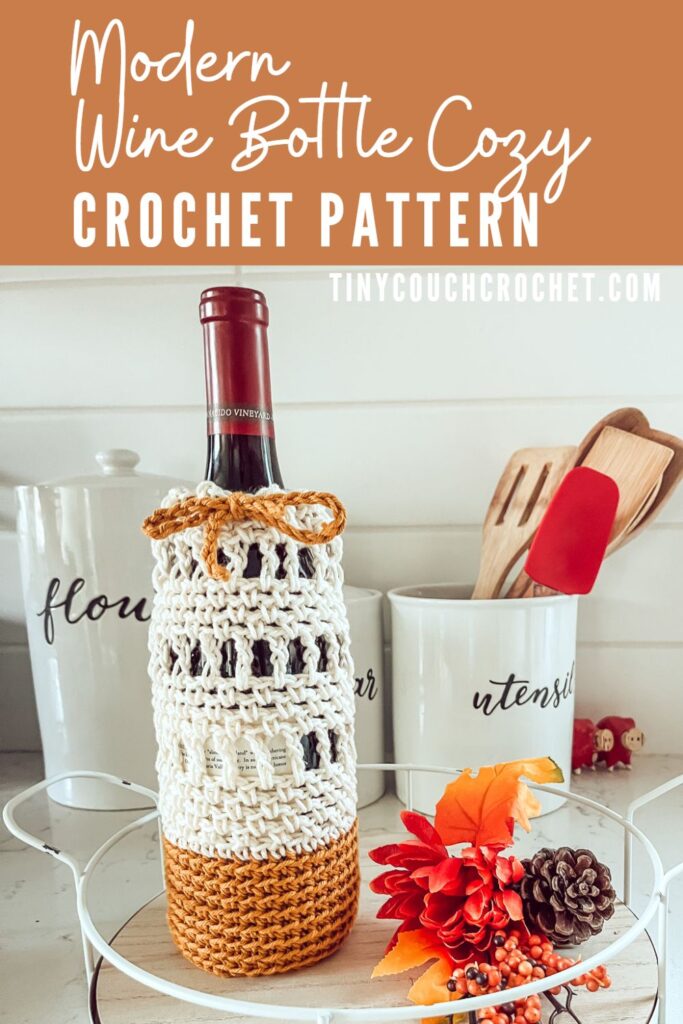 White text on a burnt orange background says "Modern wine bottle cozy crochet pattern", the bottom of the pin is an image of a wine bottle with a cream and burnt orange crochet wine bottle cozy with a drawstring to cinch the top. The wine bottle is sitting on a counter top with other kitchen and autumn decor.