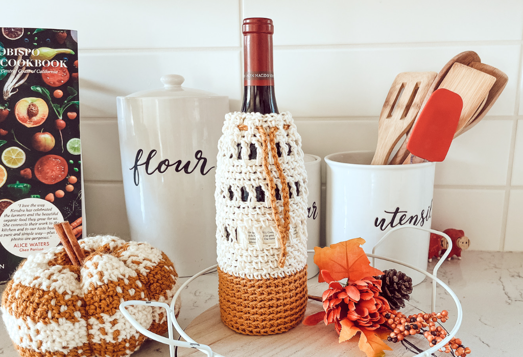 a wine bottle with a cream and burnt orange crochet wine bottle cozy with a drawstring to cinch the top. The wine bottle is sitting on a counter top with other kitchen and autumn decor.