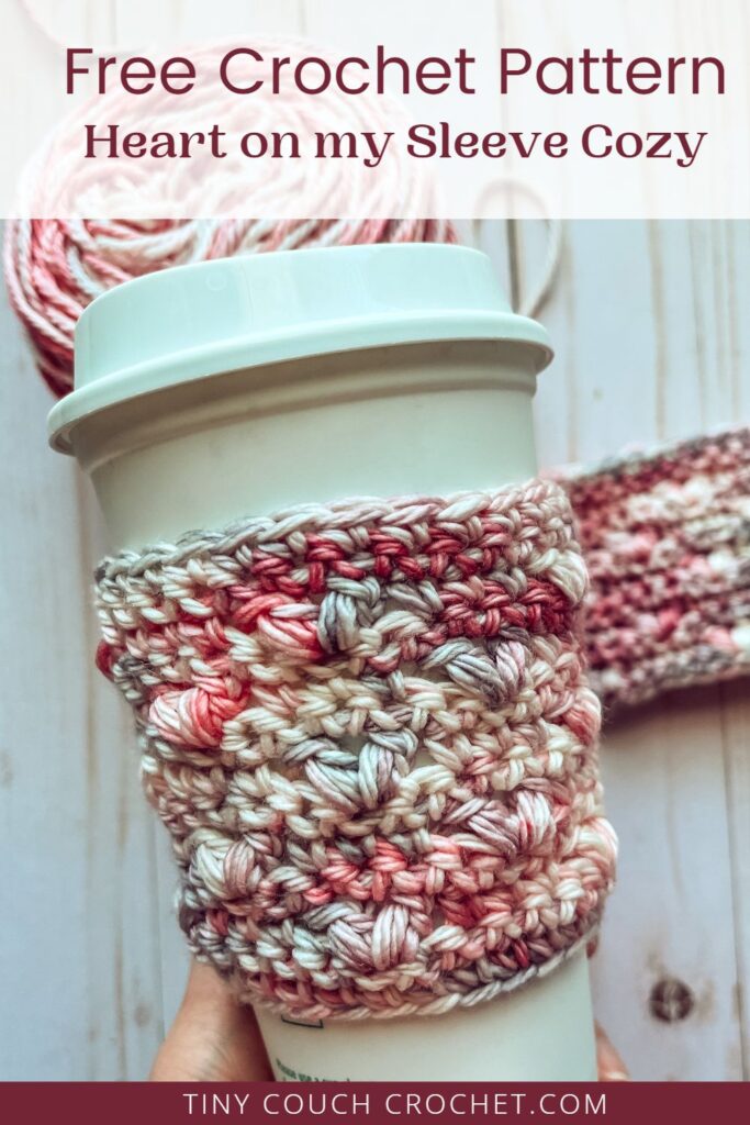 A white to-go coffee cup with a crochet cup cozy made from a free crochet cozy pattern is seen with another crochet cozy laid flat next to the yarn that was used in the background. Text at the top says "Free Crochet Pattern Heart on my Sleeve Cozy" at the top, and text that says "TinyCouchCrochet.com" at the bottom