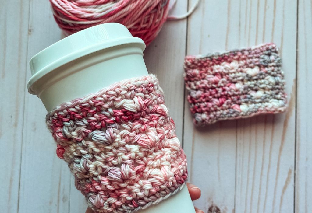 A white to-go coffee cup with a crochet cup cozy made from a free crochet cozy pattern is seen with another crochet cozy laid flat next to the yarn that was used in the background