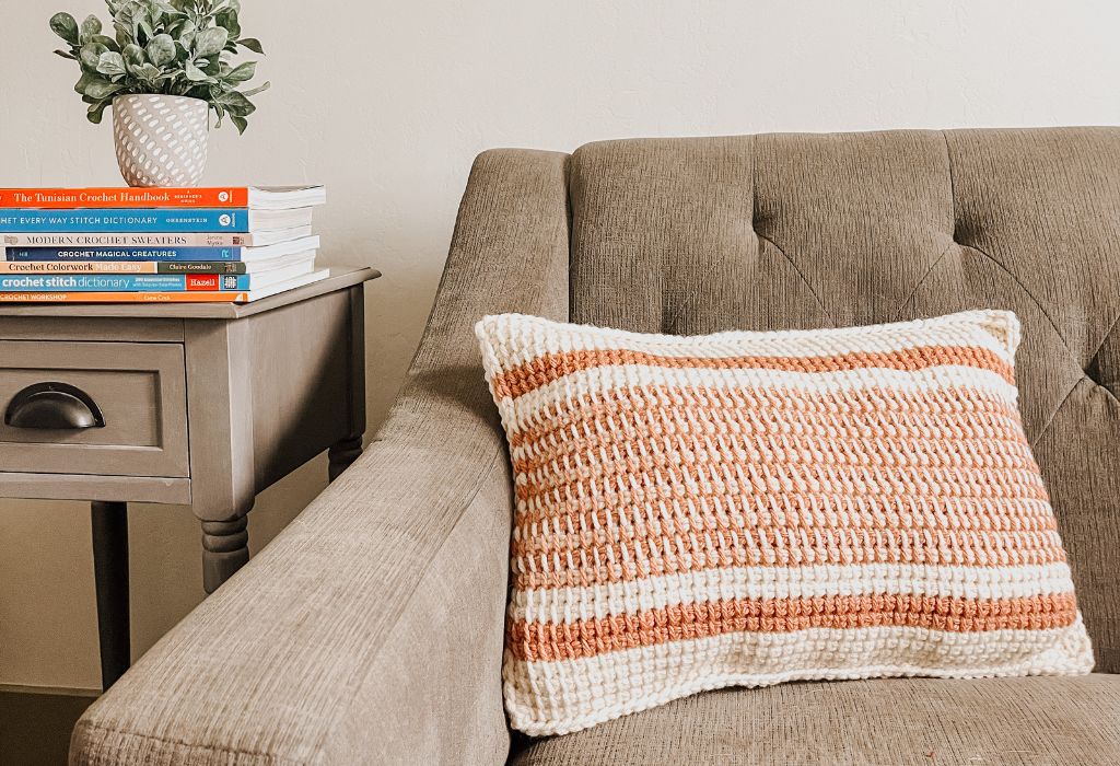 A white and pink striped tunisian crochet pillow is on a gray couch with a gray side table next to it. some crochet books are stacked on the side table.
